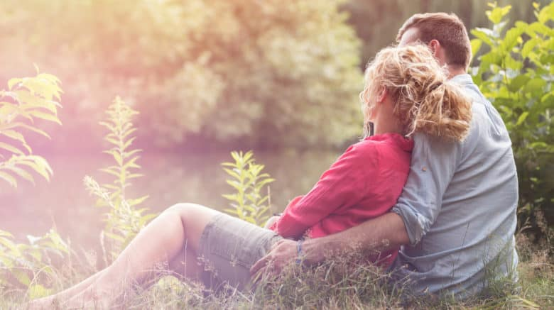 A man sits in a field of long grass surrounded by trees with a woman who is affectionately leaning her back against his chest and resting her face on his shoulder.