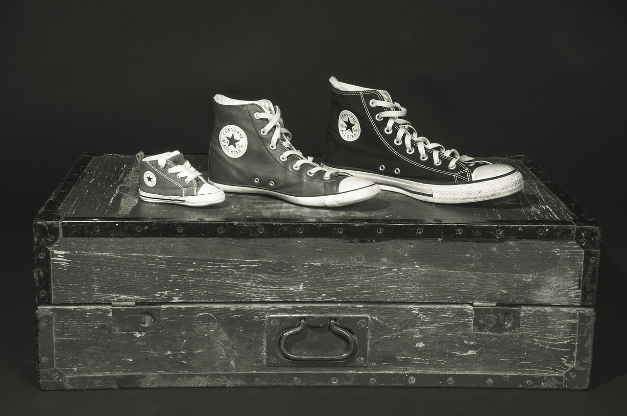 Three shoes, one very small, one medium sized, and one large, sit atop a wooden chest in a black-and-white photograph.