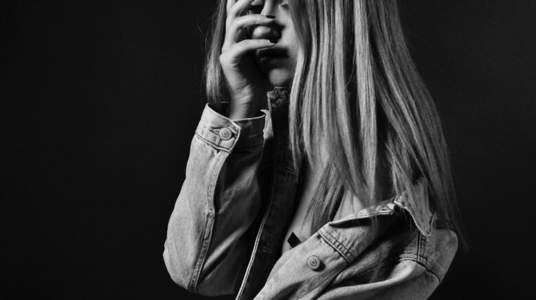 A young woman sits with her side to the camera holding her hand up to cover her face. The image is greyscale and set against a black background in a greyscale photograph.